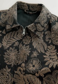 Veste noire avec des motifs floraux et cachemire beiges complexes et une fermeture éclair argentée au niveau du col avant.