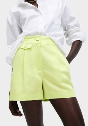 Limegrøn skræddersyede shorts med plisseringer, sidelommer og knapdetaljer, parret med en hvid skjorte med knapper. Glat stofstruktur.