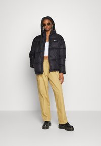 Black puffer jacket with a zip front and high collar, worn over a white tank top. Paired with beige cargo pants and black platform shoes.