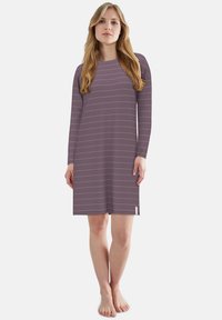 Woman standing barefoot wearing a long-sleeve, knee-length purple dress with thin horizontal pink stripes.