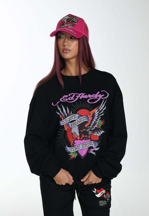 Black sweatshirt featuring colorful graphic design with eagle, heart, and text. Paired with a pink embroidered cap.