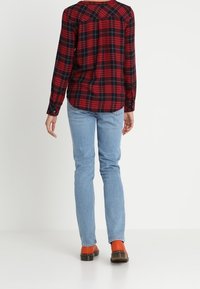 Chemise à carreaux rouge et noir à manches longues, jean bleu clair à jambes droites, chaussettes orange et chaussures marron. Modèle vu de dos.