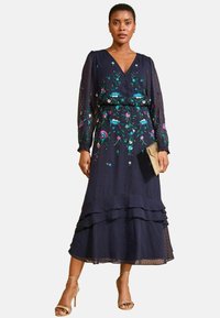 Temno modra obleka do kolen z rožnatim vezenjem, dolge prosojne rokave, v-izrez in nabrano spodnje rob. V kombinaciji z majhno zlato torbico.