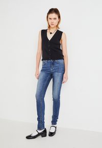 Black sleeveless button-up top paired with slim-fit blue jeans. Model wears black patent leather shoes with white socks. Plain background.