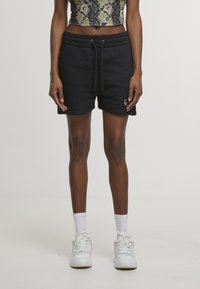 Vékony személy fekete zsinóros rövidnadrágban, fehér zokniban, fehér sportcipőben és egy rövidített kígyómintás felsőben, egyszínű háttér előtt áll.