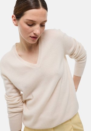 Femme à la peau claire et aux cheveux bruns, portant un pull beige à col en V et un pantalon jaune, regardant vers le bas avec sa main droite sur la hanche.