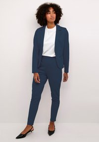 Tamsiai mėlynas kostiumas su pritaikytu švarku ir siaurais šortais, derinamas su balta T-shirt ir juodomis slingback pakulnėmis. Lygus audinys, švarios linijos.
