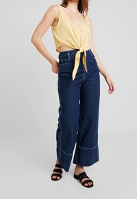 Gelbes, kurzes Tanktop mit Knöpfen vorne, das an der Taille gebunden ist. Dazu kombiniert eine hoch taillierte, weit geschnittene dunkelblaue Jeans mit Kontrastnähten.