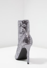 Botín de tobillo plateado con lentejuelas, tacón alto y punta redonda, textura suave y borde superior elástico para facilitar su uso. Suela negra.