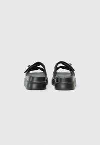 Black sandals with a textured strap, platform sole, and gold hardware accents, showing a rear view and flat, minimalist design.