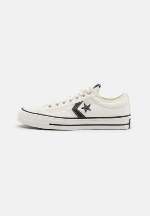 Baskets blanches en toile basse avec un logo étoile noire et un chevron, des lacets blancs et une semelle en caoutchouc rayée noire.