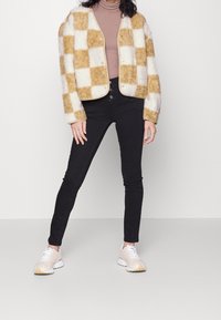 Fuzzy jacka med ett beige och krämfärgat rutmönster, rundad halsringning och framsnap-lås, kombinerad med svarta slim-fit jeans och ljusa sneakers.