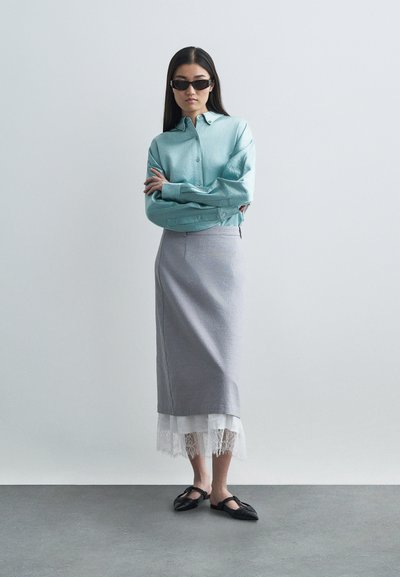 Mėlynos spalvos marškiniai su ilgomis rankovėmis, pilka sijonas su nėriniu apačioje ir juodi plokšti bateliai, prieš neutralų foną.