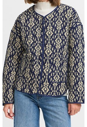 Frau trägt eine marineblaue und beige gemusterte Jacke mit Knöpfen über einem weißen Rollkragenpullover und blauen Jeans, steht mit entspannt an den Seiten hängenden Händen.