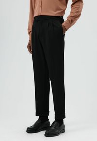 Zwarte op maat gemaakte pantalon met plooien, omgeslagen zomen en zijzakken. Draagt zwart loafers en een roze shirt met lange mouwen.