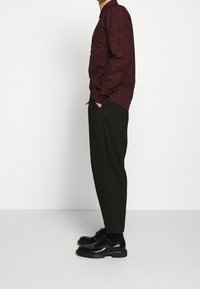 Camicia a maniche lunghe a motivi rossi abbinata a pantaloni neri e scarpe nere. La camicia presenta un colletto con bottoni e polsini visibili.