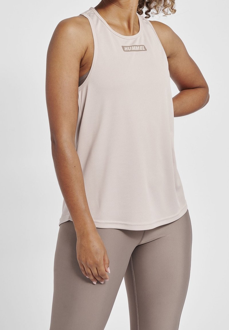 Canotta da allenamento senza maniche di colore rosa chiaro con una finitura texturizzata e un piccolo logo "HUMMEL" sul petto. Abbinata a leggings beige.