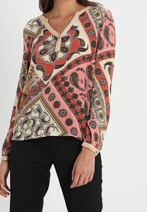 Femme portant un chemisier à manches longues avec des motifs patchwork paisley multicolores et floraux, associé à un pantalon noir.