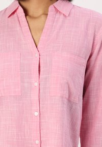 Pink checkered button-up shirt with a collar, featuring two chest pockets and a lightweight, textured fabric.