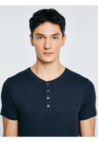 Joven con cabello corto y rizado de color negro, vistiendo una camiseta henley de manga corta azul marino, mirando hacia adelante con expresión neutral.