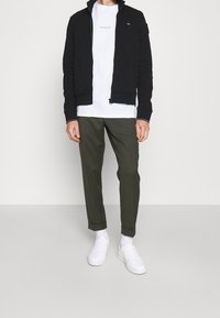 Man som bär vita sneakers, vita strumpor, uppvikta olivgröna byxor, vit t-shirt och svart zip-up jacka, stående mot en vit vägg.