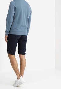 Felpa azzurra con polsini a costine, abbinata a pantaloni corti blu navy. Il modello indossa scarpe da ginnastica bianche. Vista da dietro.
