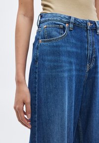 Τζιν παντελόνια με φαρδύ μπατζάκι σε μπλε χρώμα και ψηλή μέση, με χαλκοχάλκινα εξαρτήματα, μπροστινές τσέπες και ελαφρύ βαθμιαίο χρωματισμό από σκούρο σε ανοιχτό.