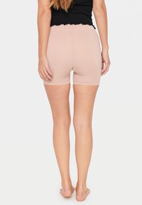 Smal kvinna som står barfota vänd bort, iförd ljusrosa tajta shorts och en svart ärmlös topp mot en enkel vit bakgrund.