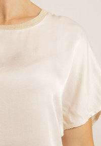 Haut blanc à manches courtes et encolure ronde. Le col présente une subtile bordure dorée. Le tissu semble doux et légèrement texturé.