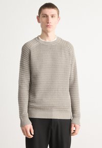Grauer Strickpullover mit strukturiertem Muster, runder Halsöffnung und gerippten Bündchen, getragen über dunklen Hosen vor einem hellen Hintergrund.