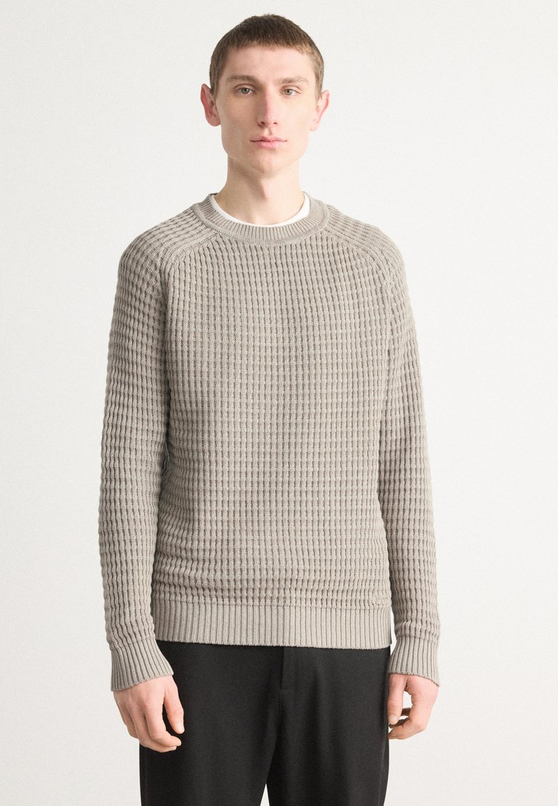 Grauer Strickpullover mit strukturiertem Muster, runder Halsöffnung und gerippten Bündchen, getragen über dunklen Hosen vor einem hellen Hintergrund.