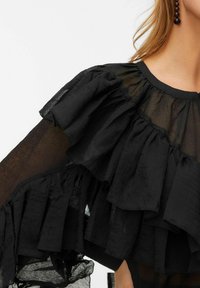 Černý top s průhlednou tkaninou, vrstvenými volány podél rukávů a výstřihu, složitou texturou a kulatým límcem.