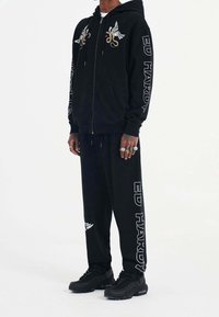 Svart zip-up hoodie med broderier av vingar och en orm, matchas med byxor i samma stil som har "ED HARDY"-text. Texturerad tyg och avslappnad passform.