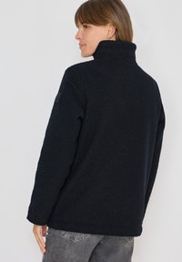 Sort fleece pullover med høj krave, struktureret overflade og diskret branding på ærmet. Enkel, lige skæring.