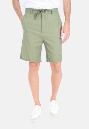 Uomo che indossa pantaloncini verde oliva con coulisse, sneakers bianche e camicia bianca, in piedi con una mano in tasca su sfondo bianco.