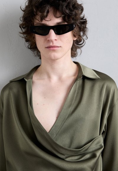 Olivengrünes Satin-Shirt mit tiefem V-Ausschnitt, lässiger Passform und drapiertem Detail an der Brust, kombiniert mit kantigen schwarzen Sonnenbrillen.
