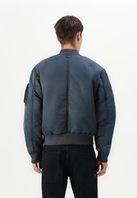Giacca bomber blu navy con orlo aderente, colletto rotondo e polsini a costine. Presenta una tasca con zip sulla manica sinistra e una finitura opaca.