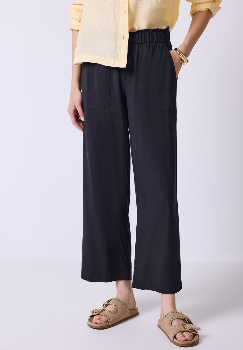 Black wide-leg pants with an elastic waistband, two front pockets, and a smooth texture, paired with beige slip-on sandals.