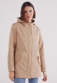 Giacca impermeabile beige con cappuccio, due tasche anteriori, taglio dritto e chiusura a bottoni. Tessuto liscio con dettagli minimi.