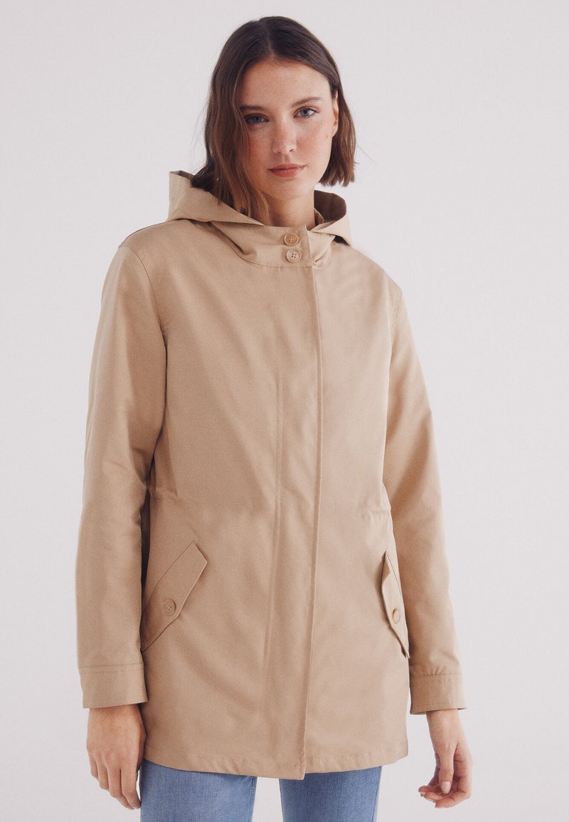 Giacca impermeabile beige con cappuccio, due tasche anteriori, taglio dritto e chiusura a bottoni. Tessuto liscio con dettagli minimi.