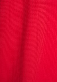 Tessuto rosso strutturato con un motivo a nido d'ape. Il materiale appare leggero e leggermente elasticizzato, adatto per abbigliamento informale o da sovrapporre.