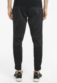 Mann trägt schwarze schmal geschnittene Hosen mit elastischen Bündchen und schwarze Puma-Sneaker, steht vor einem weißen Hintergrund.