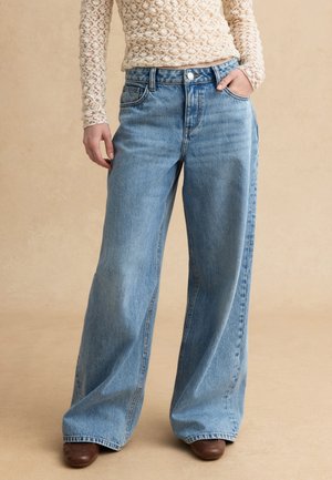 Persona con vaqueros anchos de pierna en azul claro, una blusa de encaje beige de manga larga y zapatos marrones de punta cuadrada, con la mano en el bolsillo sobre un fondo liso.