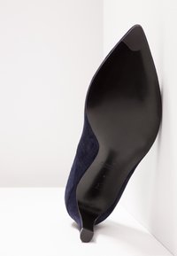 Chaussure à talon haut en daim bleu marine avec un bout pointu, une silhouette élégante et une semelle noire lisse ; design minimaliste sans ornements.