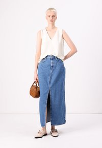 Denim maxi szoknya elől hasítékkal, világoskék színben, egy ujjatlan krémszínű toppal és egy barna fonott táskával párosítva. Lapos cipők egészítik ki a megjelenést.