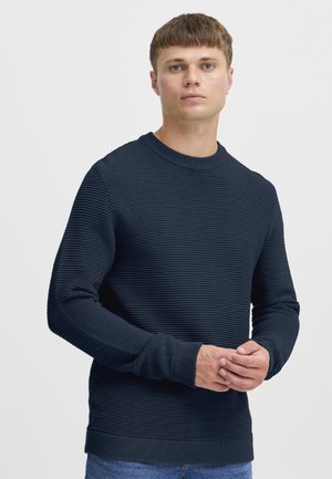 Fiatal férfi rövid barna hajjal, texturált tengerészkék hosszú ujjú pulóvert és kék farmert viselve, sima háttér előtt állva.