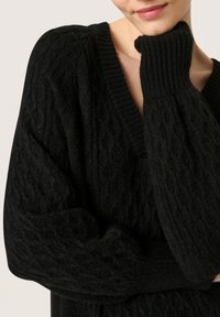 Pulover negru din tricot cu un decolteu în V, mâneci supradimensionate, manșete răsucite și un finisaj texturat. Materialul pare moale și călduros.