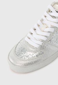 Sølvglitrende sneakers med en glatt overflate, med hvite snørebånd og rund tå. Materialet er reflekterende med en myk tekstur.