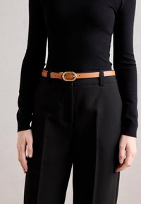 Tan leren riem met een gedraaide gouden metalen gesp, gedragen over zwarte high-waisted broek en een aansluitend zwart langemouwen shirt.