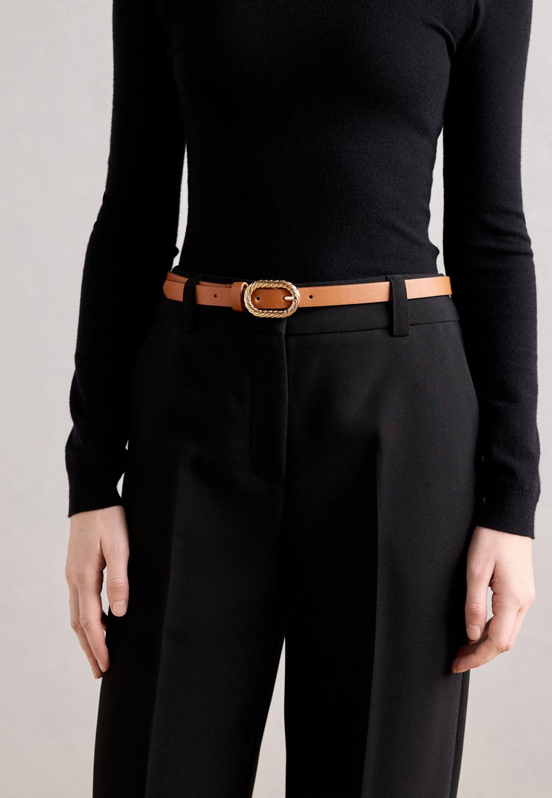 Tan leren riem met een gedraaide gouden metalen gesp, gedragen over zwarte high-waisted broek en een aansluitend zwart langemouwen shirt.